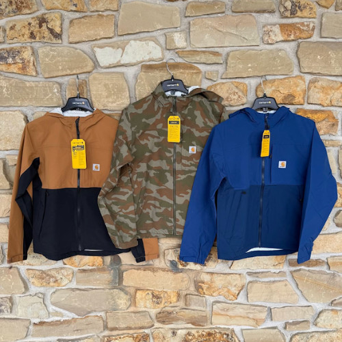 Three Carhartt jackets hang on a stone wall: tan-black, camo, and blue. Each has a hood and brand tag, with a rugged, outdoor vibe.
