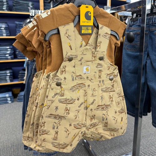 Brown toddler overalls with a nature-themed pattern hang in a store. The Carhartt tag is visible. Jeans and a stack of pants are in the background.