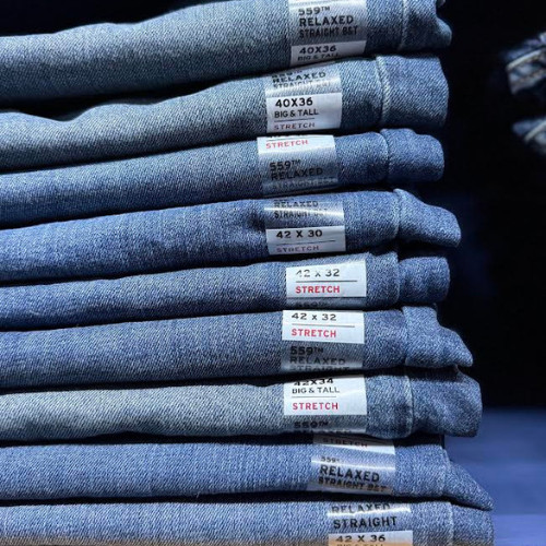 Stack of folded blue jeans with size labels visible, including various stretch fits and sizes such as 40x36 and 42x32, under soft lighting.