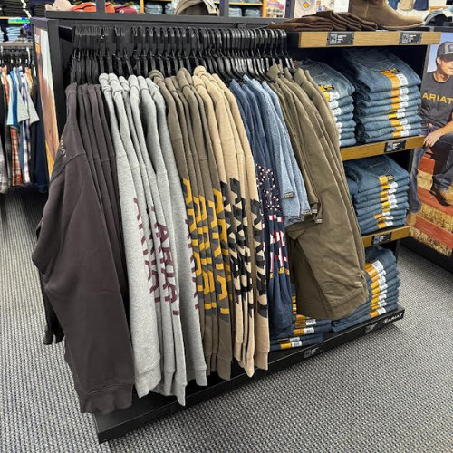 Retail clothing rack displays hoodies, sweatshirts, and jackets in gray, tan, blue, and black, with neatly folded jeans stacked behind.