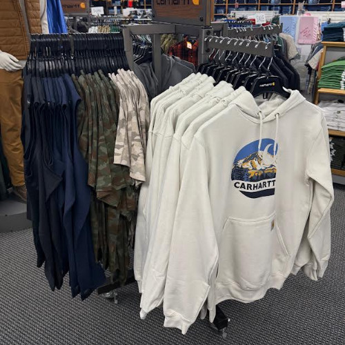 Clothing store rack displaying white Carhartt graphic hoodies, camouflage shirts, and dark jackets in a casual retail setting.