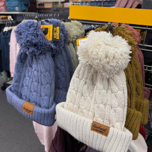 Retail display of chunky knitted winter beanies in blue, cream, gray, mustard, and burgundy, featuring fluffy pom-poms and cozy textures.