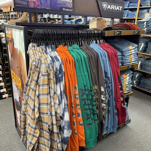 Ariat store display with colorful plaid shirts and graphic T-shirts on racks, folded jeans and apparel stacked behind in an organized retail setting.