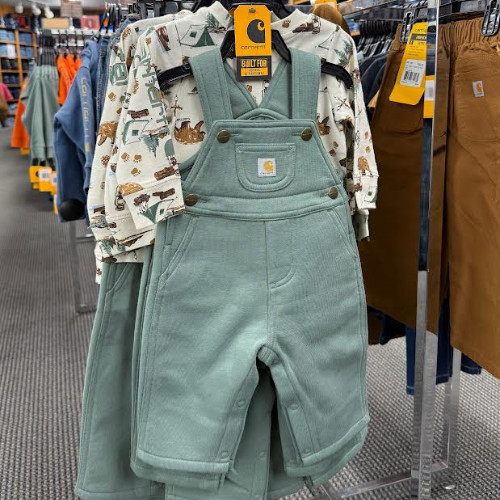 Children’s sage-green bib overalls hang over a patterned long-sleeve shirt on a retail rack, with Carhartt tags and workwear nearby.