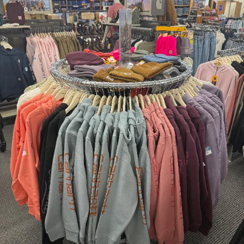 Circular clothing rack displays colorful hoodies and sweaters, with folded knitwear and busy retail aisles visible in the background.